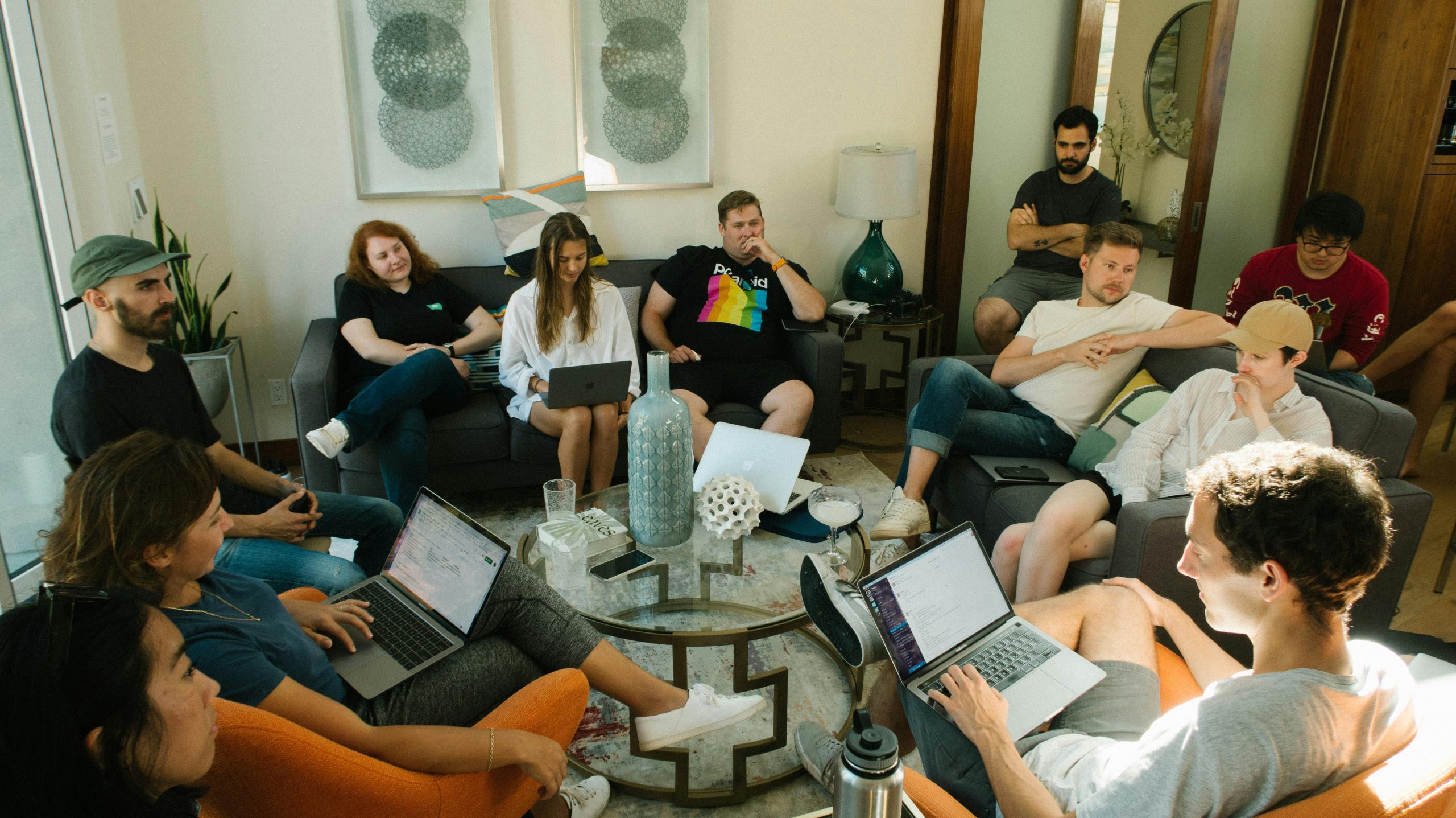 A group of people gathered in a living room for a casual meeting, sitting on couches and chairs, some with laptops, engaged in discussion.